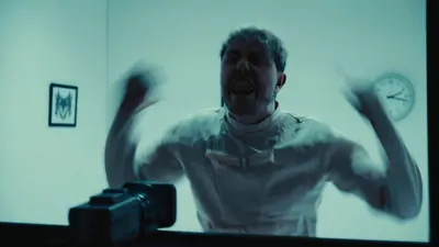 Video still: a patient in a straitjacket thrashing in a white room